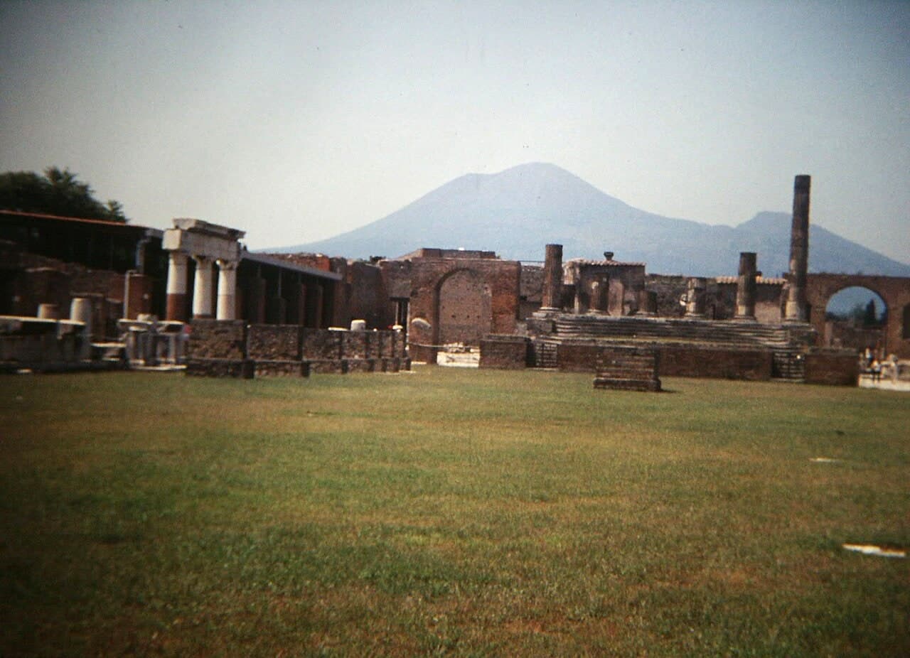 The forum at Pompeii, with a colonnade, the steps of the Temple of Jupiter, a lone column, and Mount Vesuvius behind.