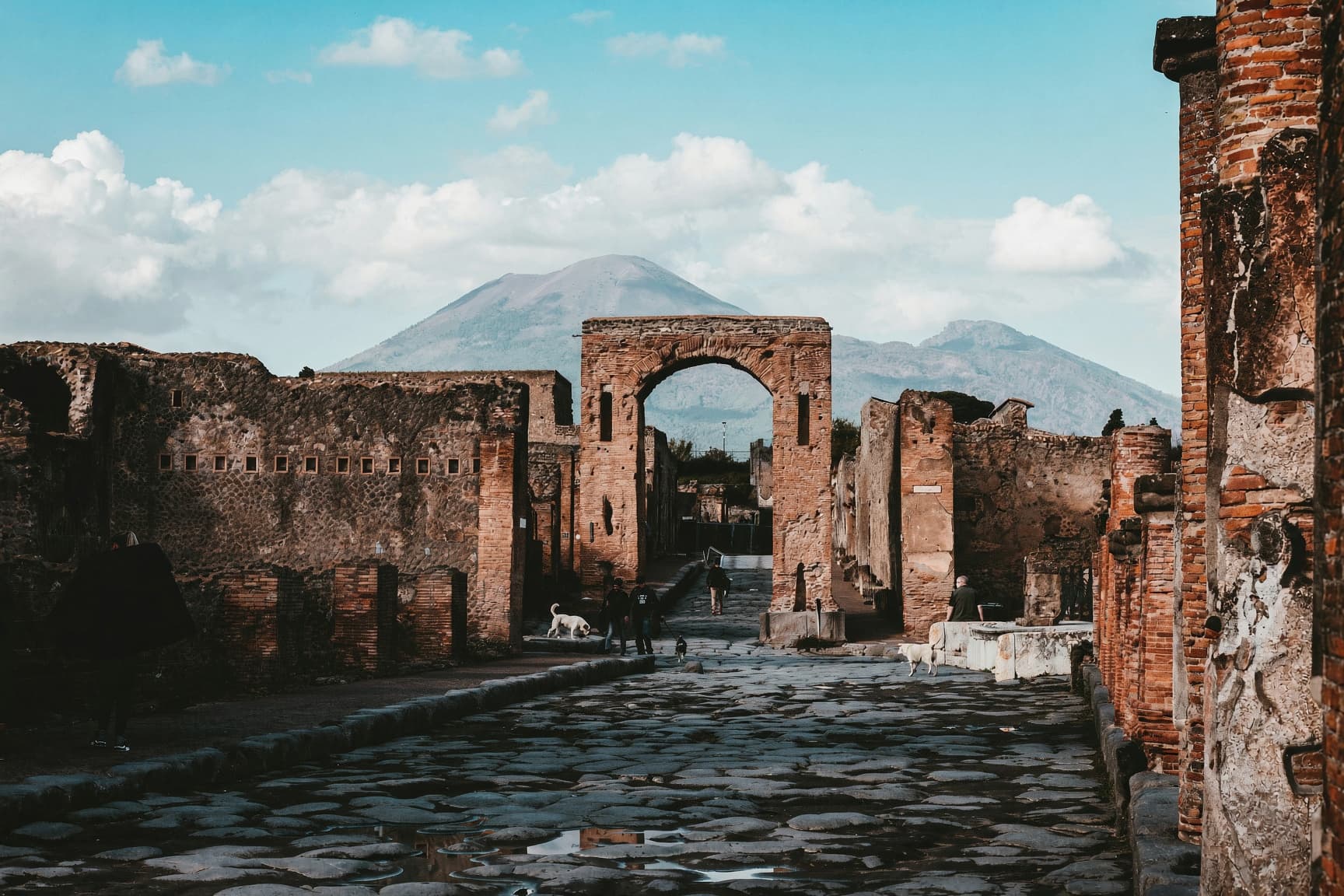 A stone street in Pompeii passing under a brick arch, with Mount Vesuvius beyond the walls.